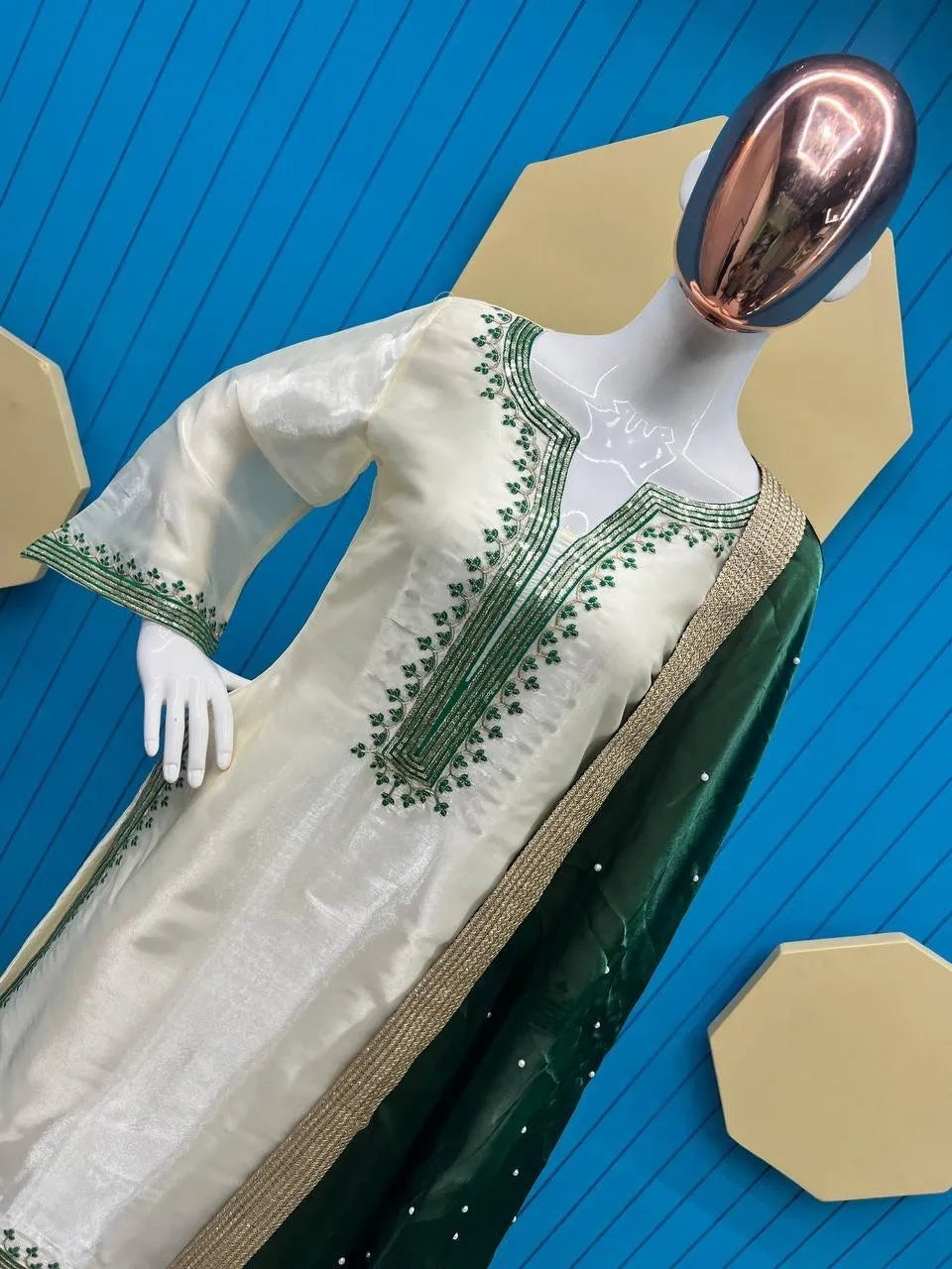 A cream-colored kurta with green embroidered details and a dark green dupatta draped over a white mannequin, set against a blue slatted background with gold geometric shapes.