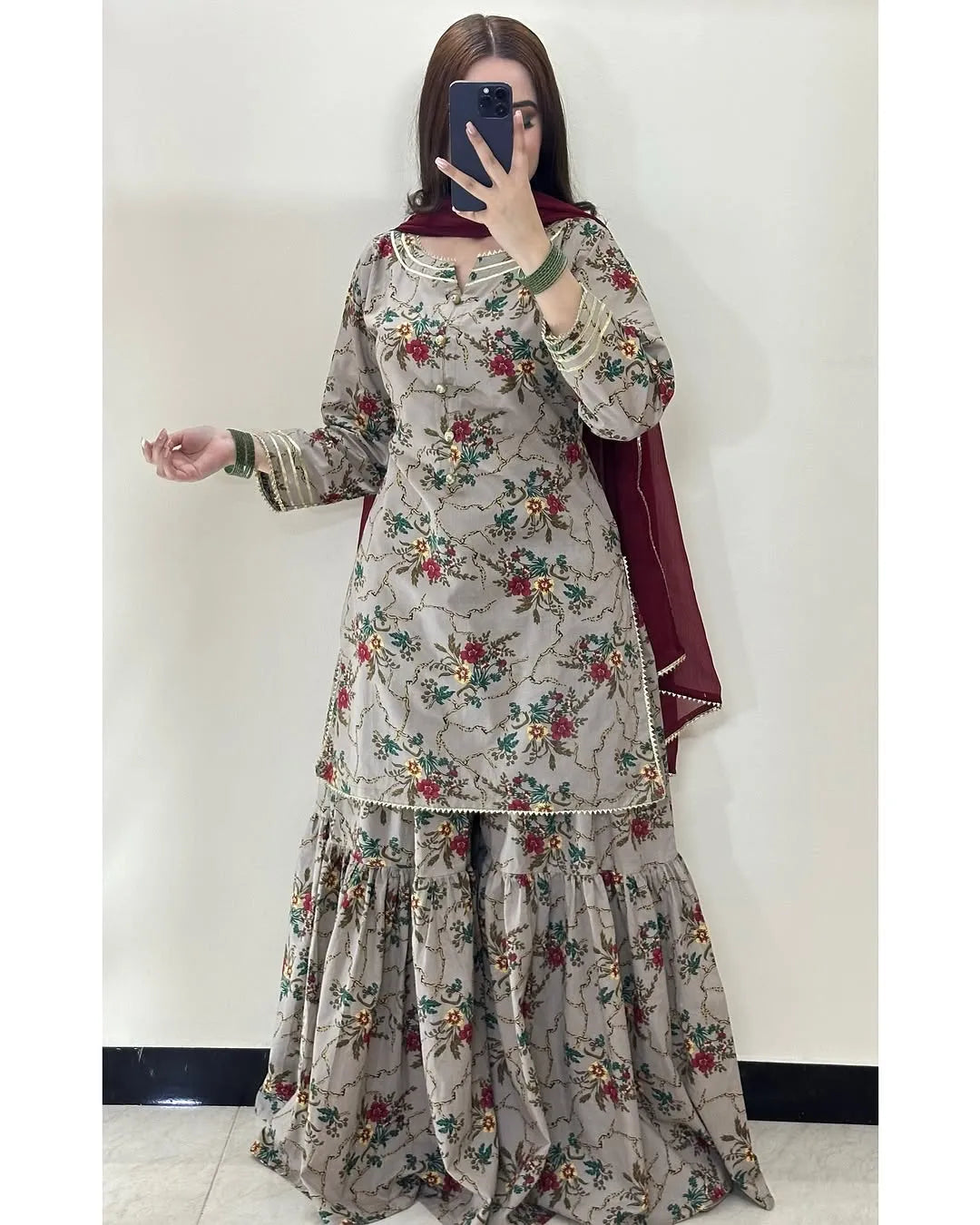 A woman takes a mirror selfie wearing a traditional three-piece salwar kameez set in beige with a red and green floral print, featuring a matching solid deep red dupatta.