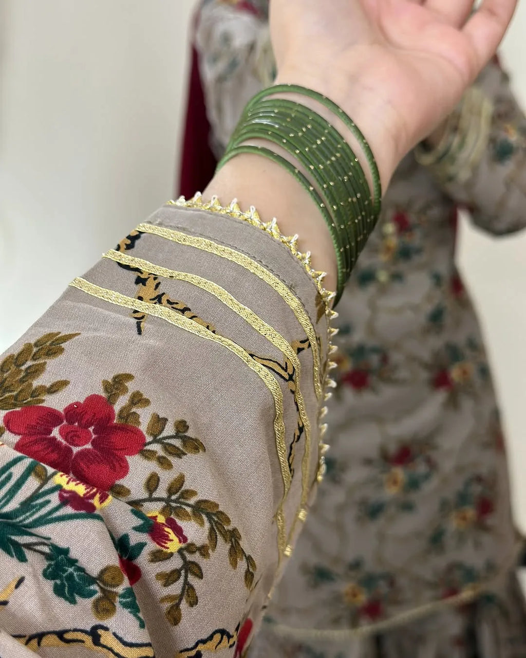 Close-up of a person's wrist wearing a stack of green beaded bangles, with a floral printed fabric sleeve featuring red flowers, green leaves, and gold trim in the foreground.