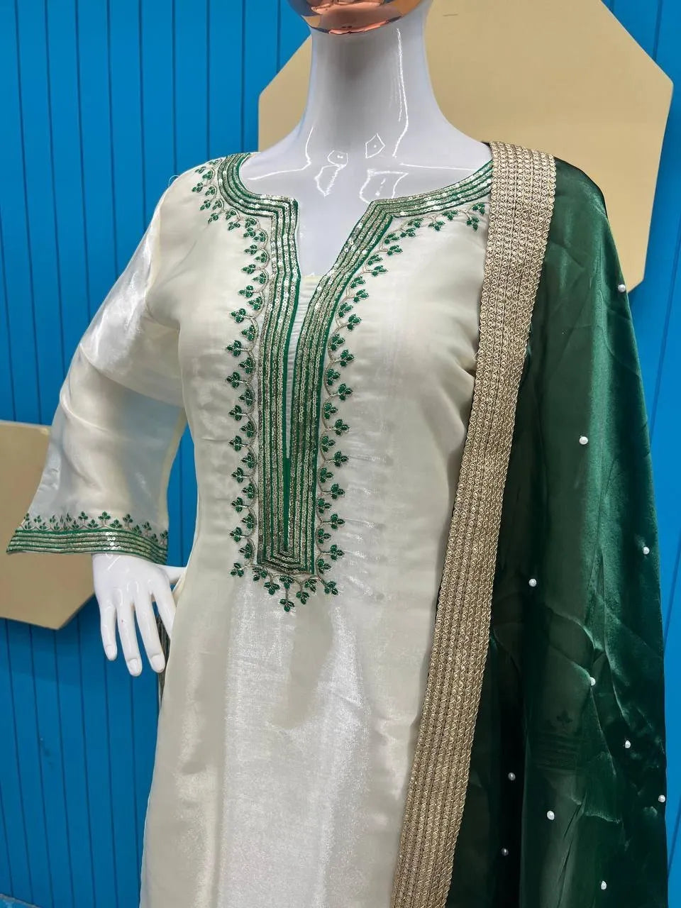 A three-piece traditional South Asian outfit displayed on a mannequin, featuring an off-white kurta with green floral embroidery, a green dupatta with silver mirror work, and a beige textured border.