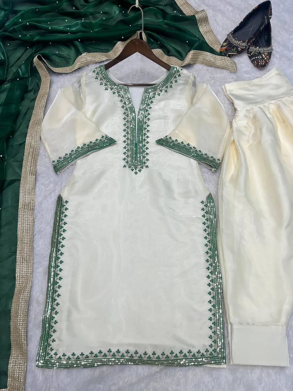 An off-white/cream traditional outfit laid flat on a light surface, featuring a kurta with green embroidered borders and matching pants. A dark green dupatta with metallic trim and a pair of embellished flats are also arranged around it.