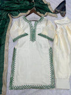 An off-white/cream traditional outfit laid flat on a light surface, featuring a kurta with green embroidered borders and matching pants. A dark green dupatta with metallic trim and a pair of embellished flats are also arranged around it.