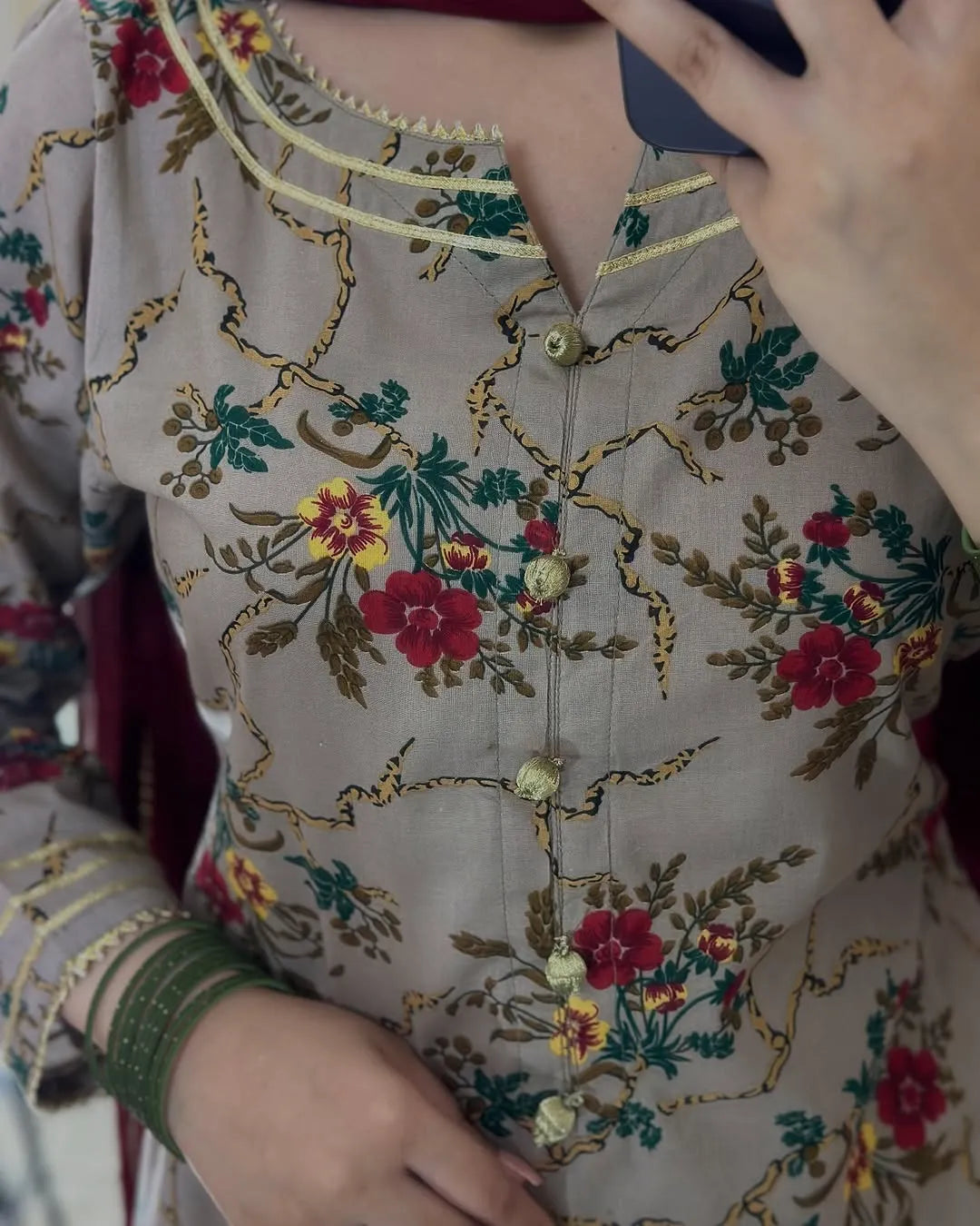 A close-up of a beige embroidered kurta featuring red and yellow floral motifs with green leaves and gold-toned decorative trim and buttons.