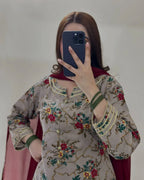 A woman takes a mirror selfie wearing a beige embroidered salwar kameez top with red and green floral patterns, green bangles, and a dark red dupatta.