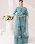 A woman models a teal blue three-piece salwar kameez suit with matching sheer dupatta. The outfit features intricate silver and gold embroidery on the chest, sleeves, and dupatta, with a drawstring neckline and wide-leg trousers.