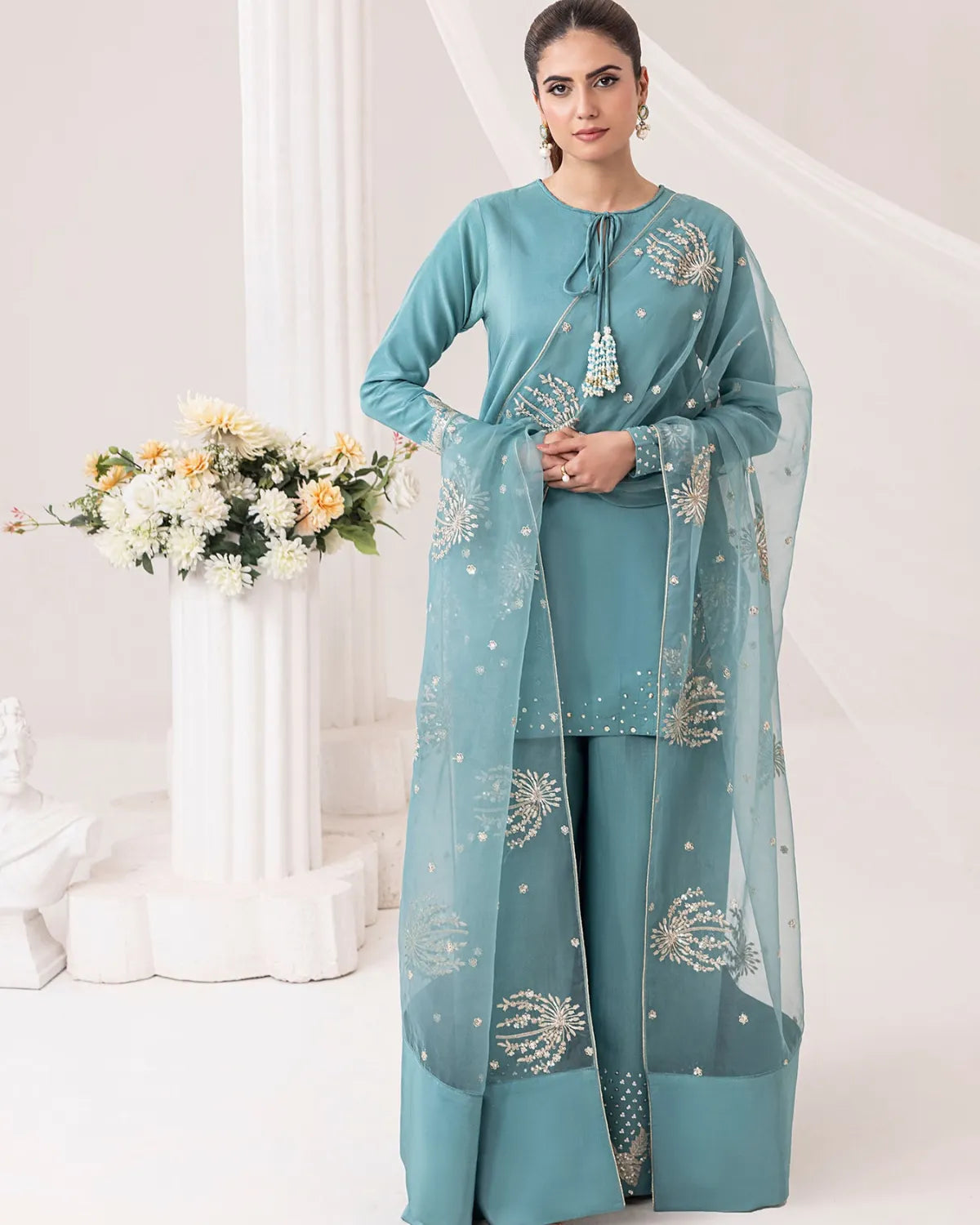 A woman models a teal blue three-piece salwar kameez suit with matching sheer dupatta. The outfit features intricate silver and gold embroidery on the chest, sleeves, and dupatta, with a drawstring neckline and wide-leg trousers.