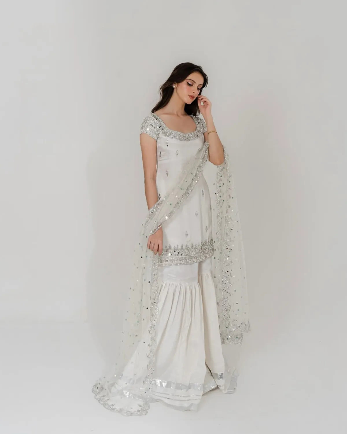 A woman modeling a white traditional outfit with silver mirror work embroidery and a matching sheer dupatta, photographed against a plain light gray studio background.
