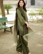 A woman wearing an olive green three-piece traditional outfit with matching sheer dupatta and embroidered sleeve details, standing outdoors in a park-like setting.