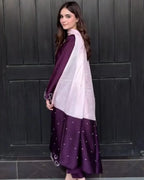 A woman wearing a traditional South Asian outfit with a deep purple skirt decorated with small silver polka dots, a matching purple dupatta, and a light lavender shawl draped over her shoulders, standing in front of a dark paneled door.