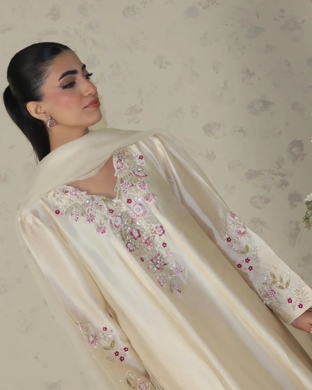 A woman wearing a cream-colored satin traditional outfit with pink and green floral embroidery, paired with a matching dupatta, set against a patterned beige wall.