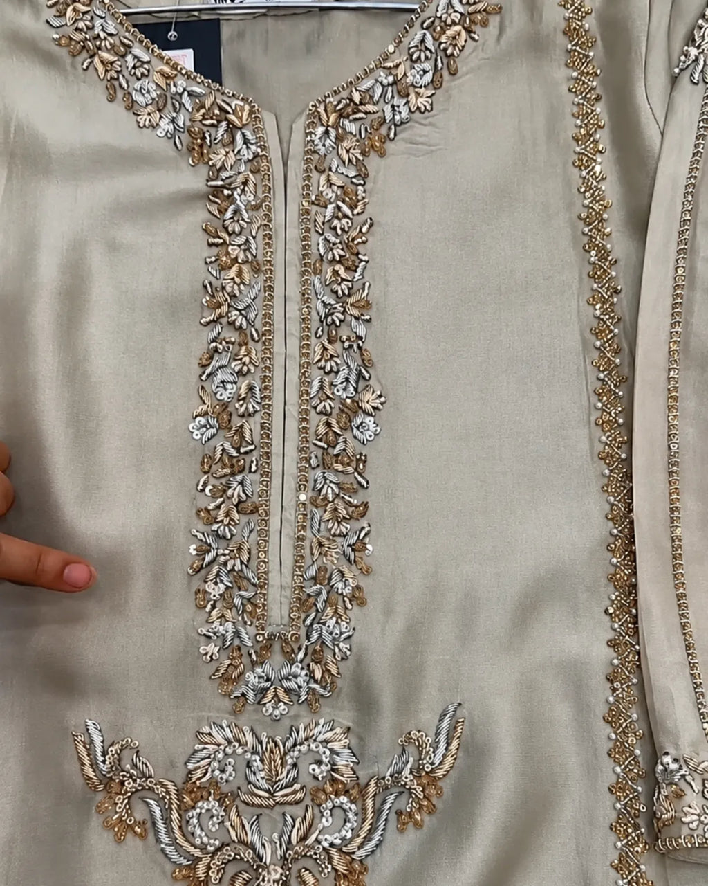 Close-up of a beige or champagne-colored embroidered garment, likely a kurta or tunic, featuring ornate metallic silver and gold floral embroidery around the neckline and lower front panel.