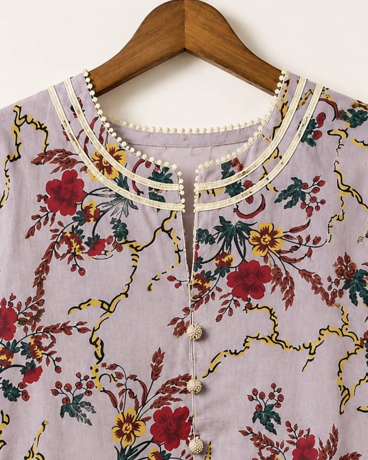 A close-up of a lavender floral printed top hanging on a wooden hanger. The top features a cream-colored collar embellished with small pearls, a matching pearl button placket, and a vibrant floral pattern with red, yellow, and green flowers on a gold vine background.