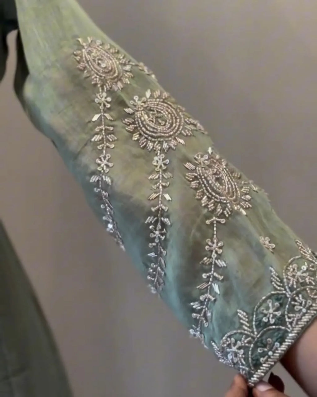 A close-up of a sage green fabric sleeve or garment piece decorated with ornate silver embroidery and beadwork, held by a hand against a plain neutral background.