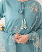 A close-up of a person wearing a light blue-green traditional outfit with silver floral embroidery and a beaded tassel tie at the neckline. The fabric is sheer, and the person's hands are clasped in front.