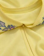Close-up of a pale yellow garment with decorative floral embroidery in blue, purple, and green along the neckline, shown against matching pale yellow fabric.