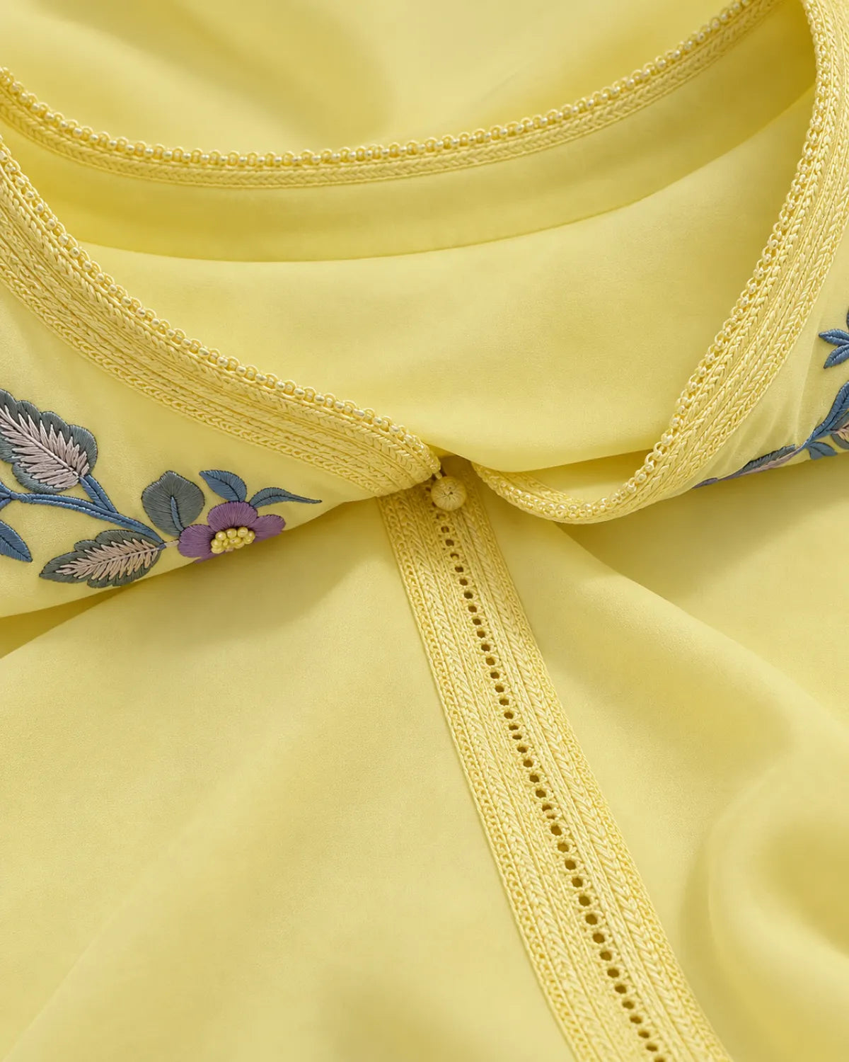 Close-up of a pale yellow garment with decorative floral embroidery in blue, purple, and green along the neckline, shown against matching pale yellow fabric.