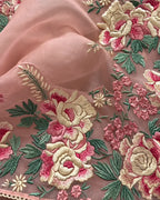 A close-up of peach-pink embroidered fabric featuring large floral motifs in cream, pink, and green, with a sheer matching fabric draped behind it.