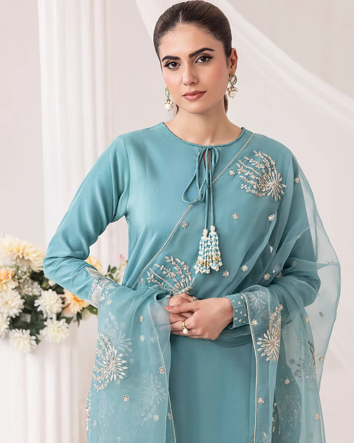 A woman modeling a light blue-green traditional outfit with matching sheer dupatta, featuring intricate silver and white floral embroidery and tassel details.