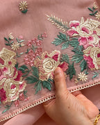 A close-up of a dusty pink embroidered fabric with large floral motifs in pink, cream, and green, held by a hand to show the texture and detail.