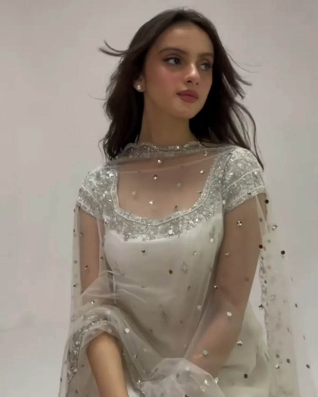 A woman wearing a white embellished traditional outfit with sheer sleeves and silver beadwork, photographed against a plain light background.