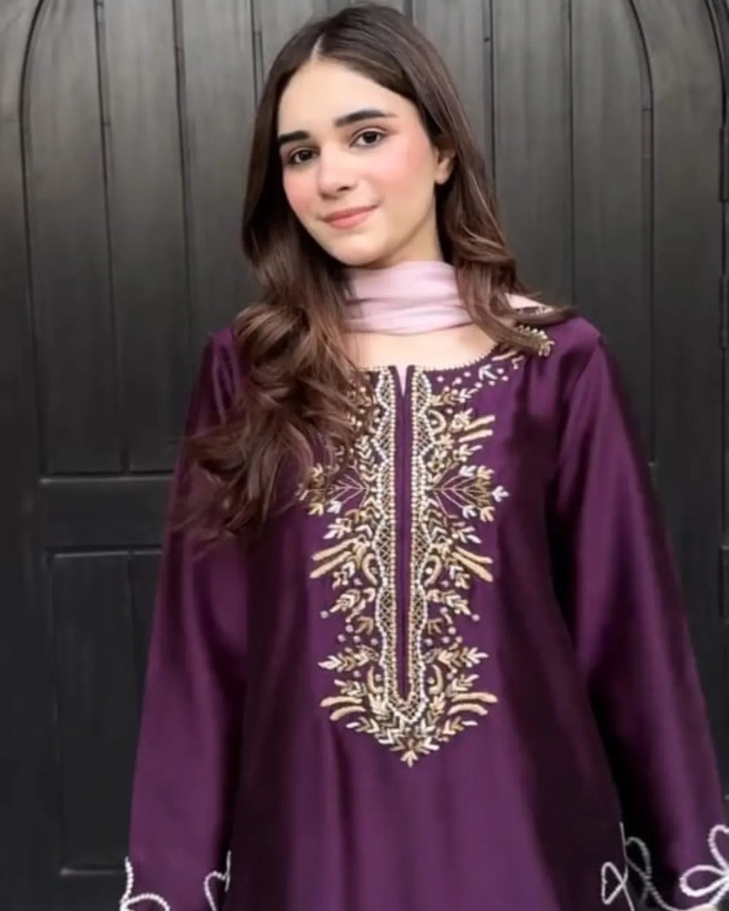 A young girl with long brown hair wearing a purple long-sleeve tunic with gold and white embroidery on the chest, paired with a light pink neck scarf, standing in front of a dark wooden door.