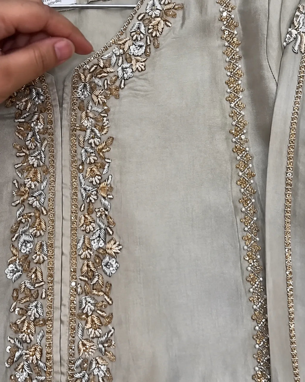 A close-up of a light beige or silver-gray fabric garment with ornate floral embroidery and beadwork in gold and silver along the neckline and front edges. A hand is visible holding the fabric at the upper left.