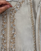 A close-up of a light beige or silver-gray fabric garment with ornate floral embroidery and beadwork in gold and silver along the neckline and front edges. A hand is visible holding the fabric at the upper left.