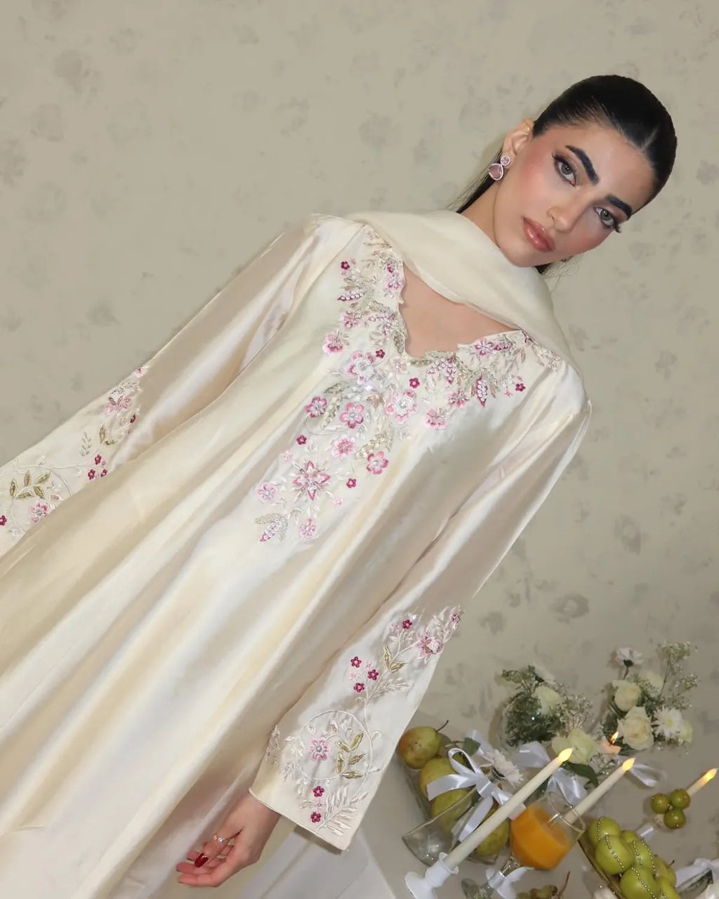 A woman wearing a cream satin abaya with pink and green floral embroidery, posed against a patterned beige wall with decorative props at the bottom right.
