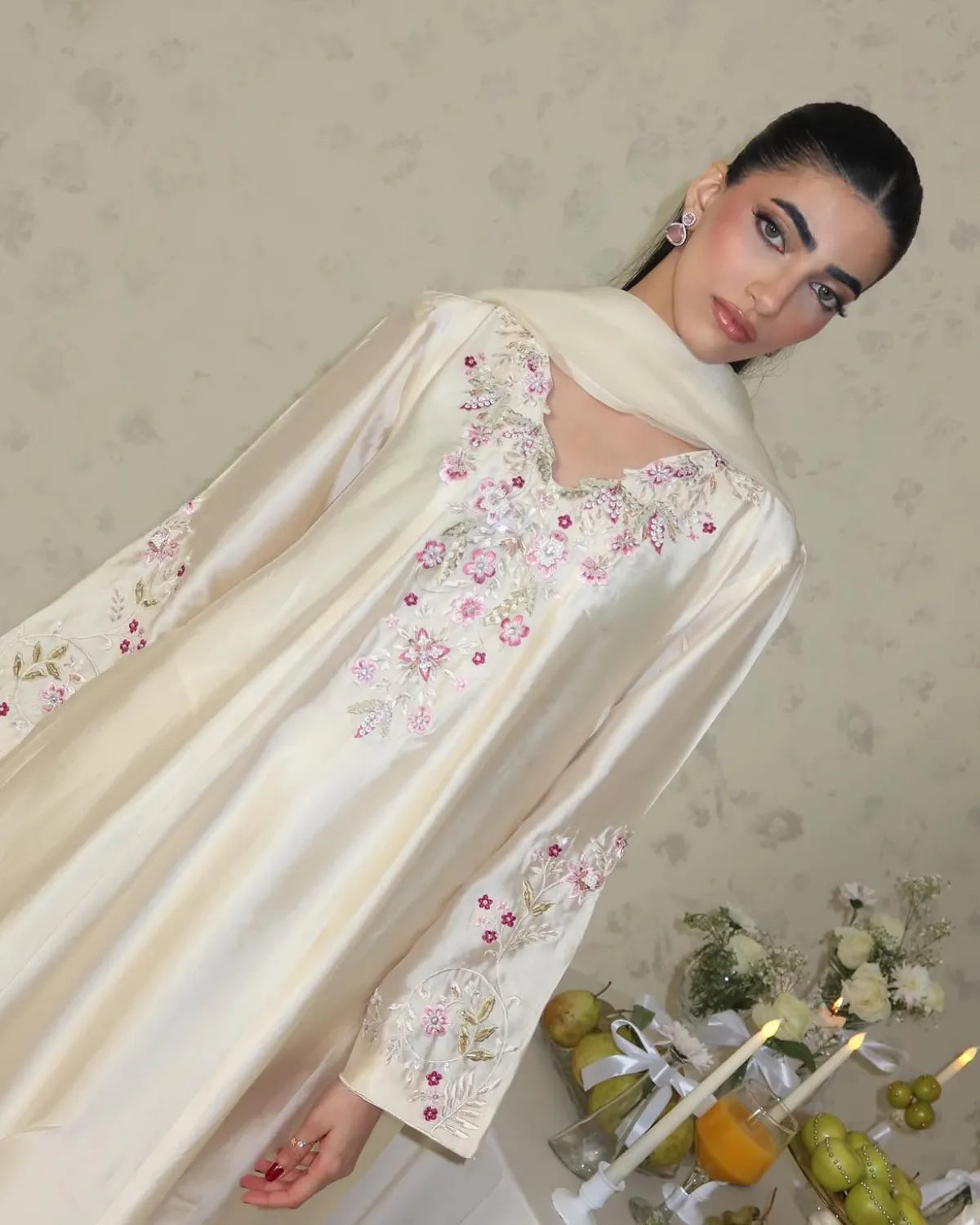 A woman wearing a cream satin abaya with pink and green floral embroidery, posed against a patterned beige wall with decorative props at the bottom right.