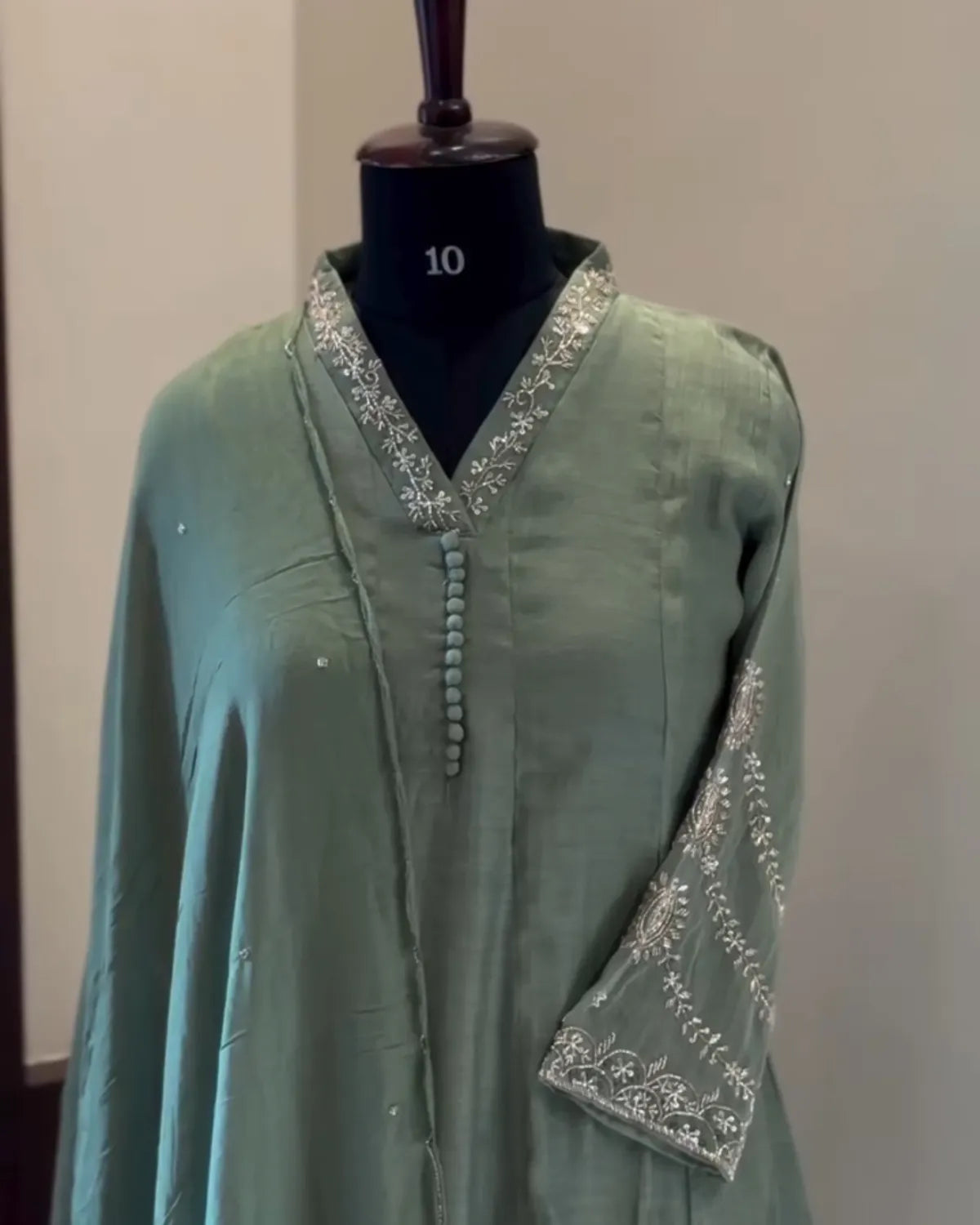 A sage green long-sleeve tunic or kurta displayed on a black mannequin torso. The garment features silver embroidered detailing along the V-neck collar, a decorative button placket, and the cuffs.