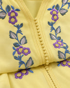 Close-up of a pale yellow garment with embroidered floral vines in purple, blue, green, and gray, plus a braided trim running down the center front.