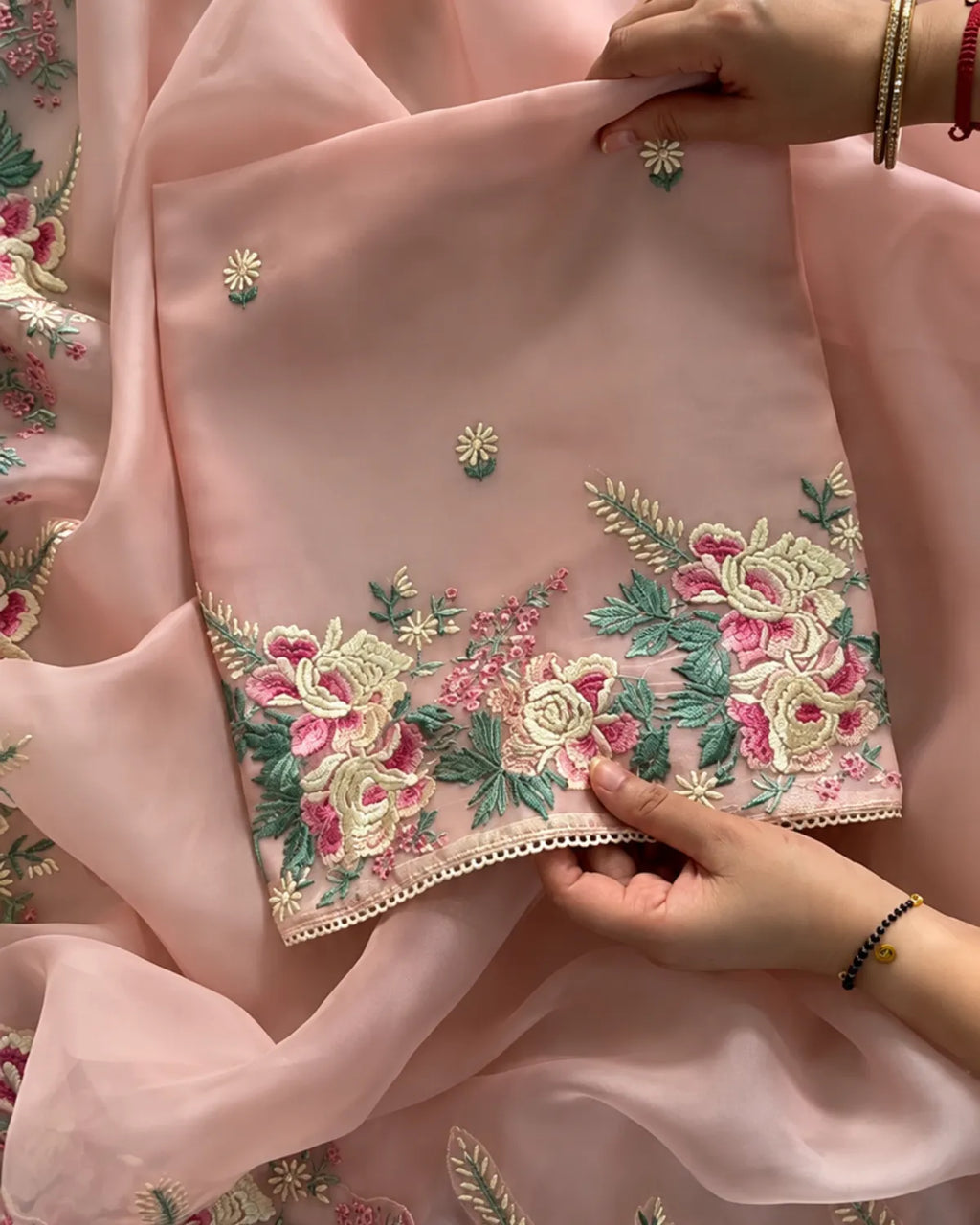 A close-up of a light pink embroidered fabric, likely a dupatta or scarf, held by hands to showcase its detailed floral embroidery. The fabric features vibrant pink and cream flowers with green leaves and small embroidered motifs, finished with a decorative scalloped border.