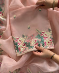 A close-up of a light pink embroidered fabric, likely a dupatta or scarf, held by hands to showcase its detailed floral embroidery. The fabric features vibrant pink and cream flowers with green leaves and small embroidered motifs, finished with a decorative scalloped border.