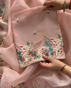 A close-up of a light pink embroidered fabric, likely a dupatta or scarf, held by hands to showcase its detailed floral embroidery. The fabric features vibrant pink and cream flowers with green leaves and small embroidered motifs, finished with a decorative scalloped border.