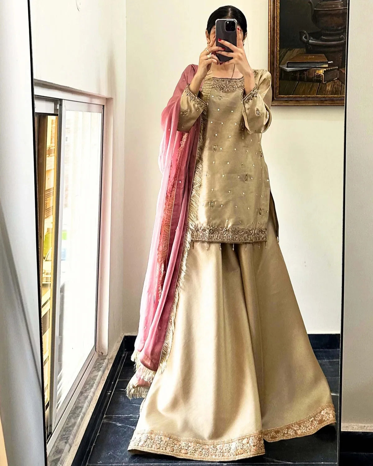 A woman takes a mirror selfie wearing a traditional beige and gold embroidered kurta set with a pink dupatta draped over her shoulder, set against an indoor background.