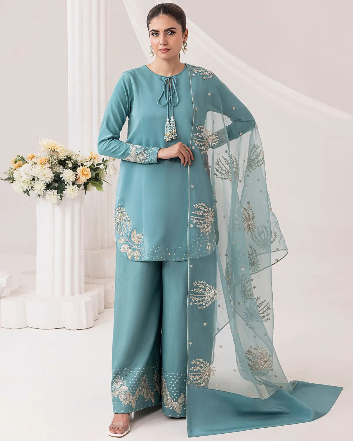 A woman models a teal blue three-piece salwar kameez suit with matching dupatta. The kurta and wide-leg trousers feature gold and silver embroidery, and the sheer dupatta has matching floral patterns.