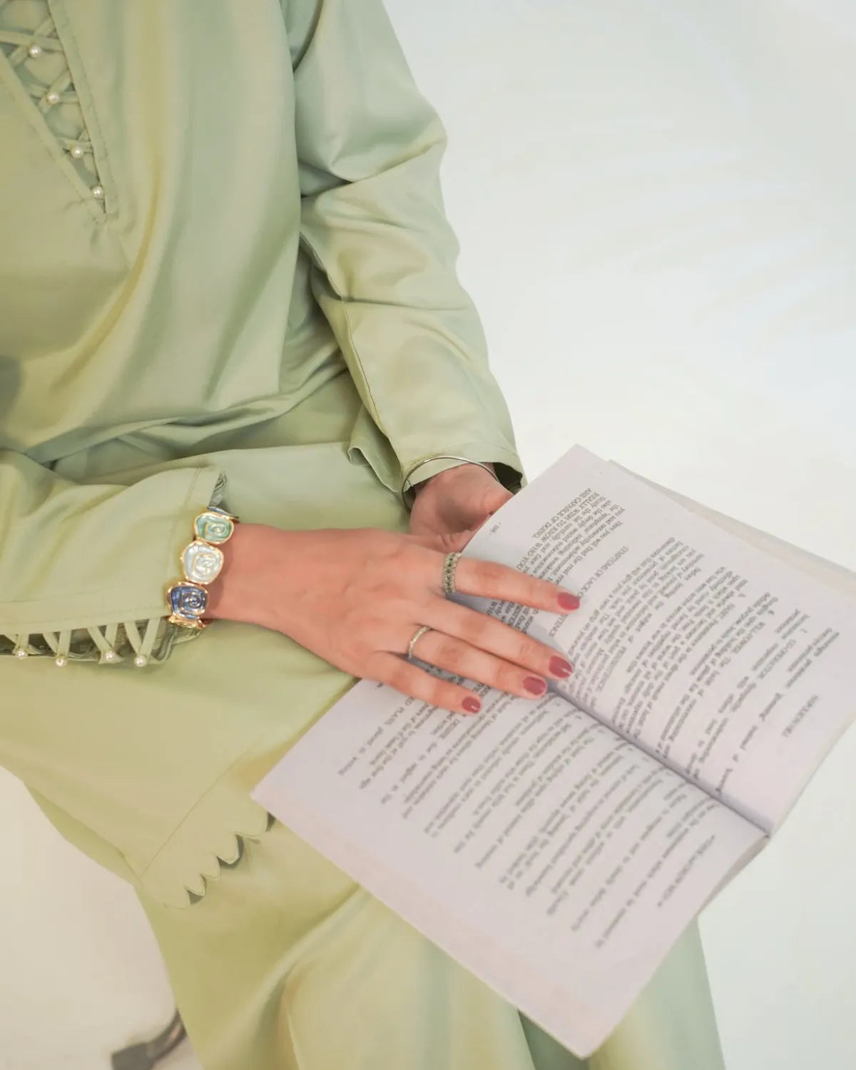 A person wearing a light sage green traditional outfit holds an open book. The outfit features decorative lace-up detailing on the chest and a scalloped edge on the sleeve.