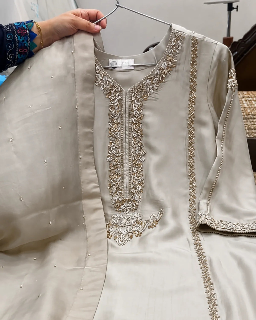 A light beige or cream-colored embroidered kurta with a matching dupatta draped over the shoulder, held up by a hand on a metal hanger. The kurta features intricate gold and silver embroidery around the neckline and along the borders.