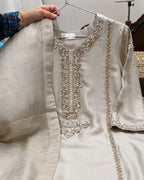 A light beige or cream-colored embroidered kurta with a matching dupatta draped over the shoulder, held up by a hand on a metal hanger. The kurta features intricate gold and silver embroidery around the neckline and along the borders.