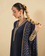 A woman wearing a navy blue traditional outfit with gold embroidery, a matching sheer dupatta, and ornate gold jewelry, posed against a plain beige background.