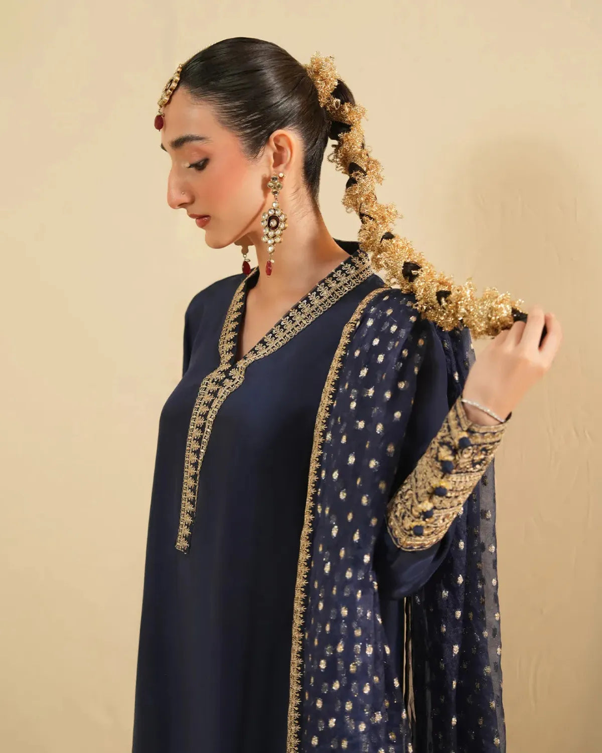 A woman wearing a navy blue traditional outfit with gold embroidery, a matching sheer dupatta, and ornate gold jewelry, posed against a plain beige background.