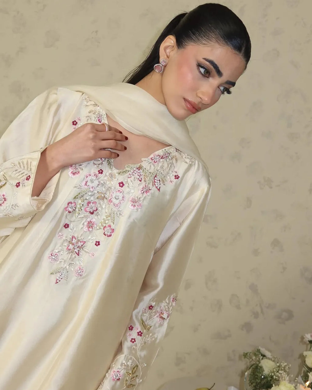 A woman wearing a cream satin kurta with pink and green floral embroidery, paired with a matching sheer dupatta, set against a patterned beige wall.