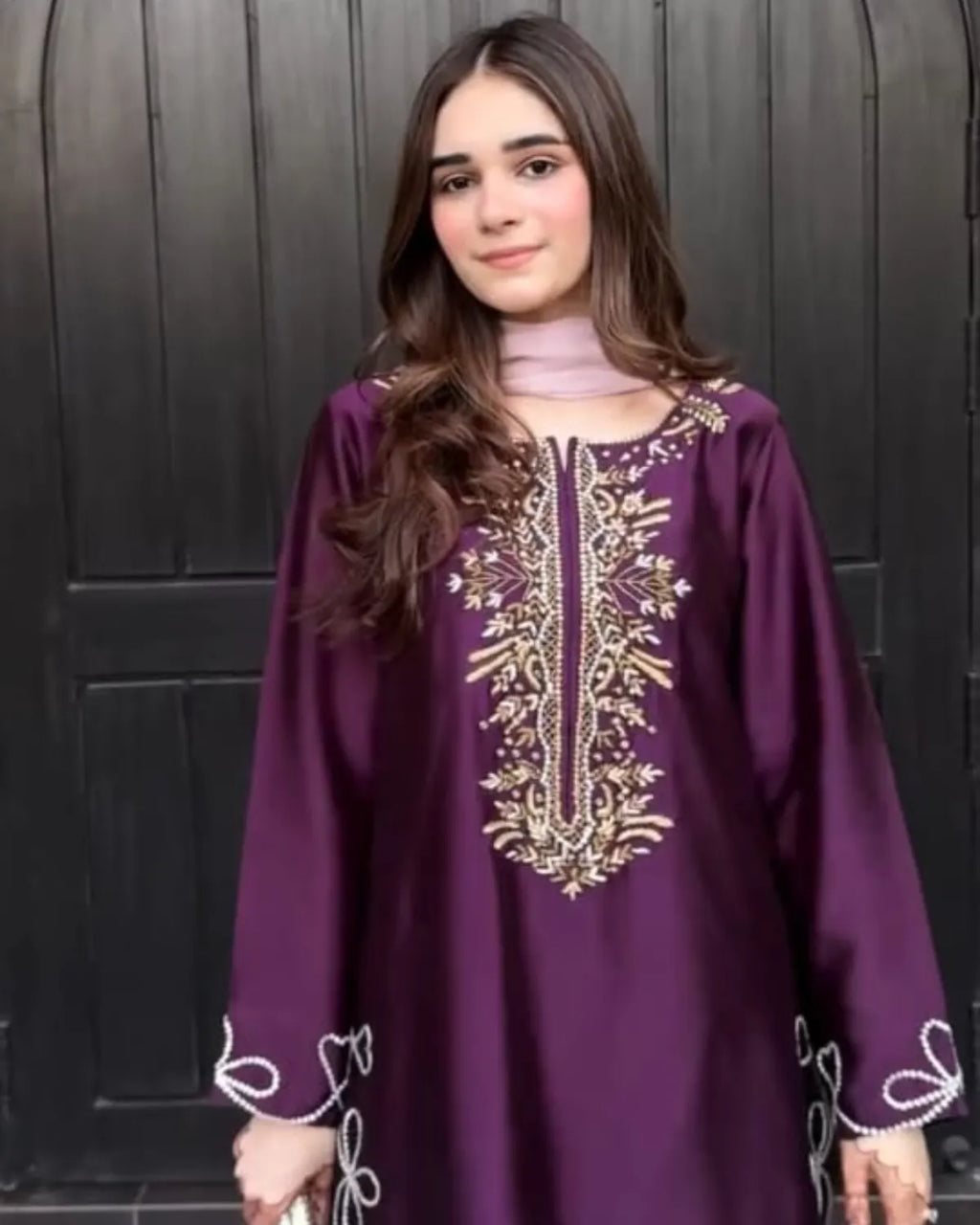 A young woman wearing a deep purple long-sleeve tunic with ornate gold embroidery on the chest and white embroidery on the cuffs, standing in front of a dark wooden door.