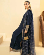 A woman models a navy blue traditional outfit with gold embroidery and a matching patterned dupatta, standing on beige steps against a cream-colored wall.