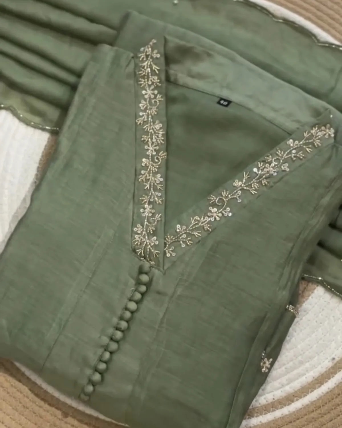An olive green fabric garment laid flat on a woven mat, featuring a V-neckline with metallic floral embroidery and a decorative row of round buttons down the front.