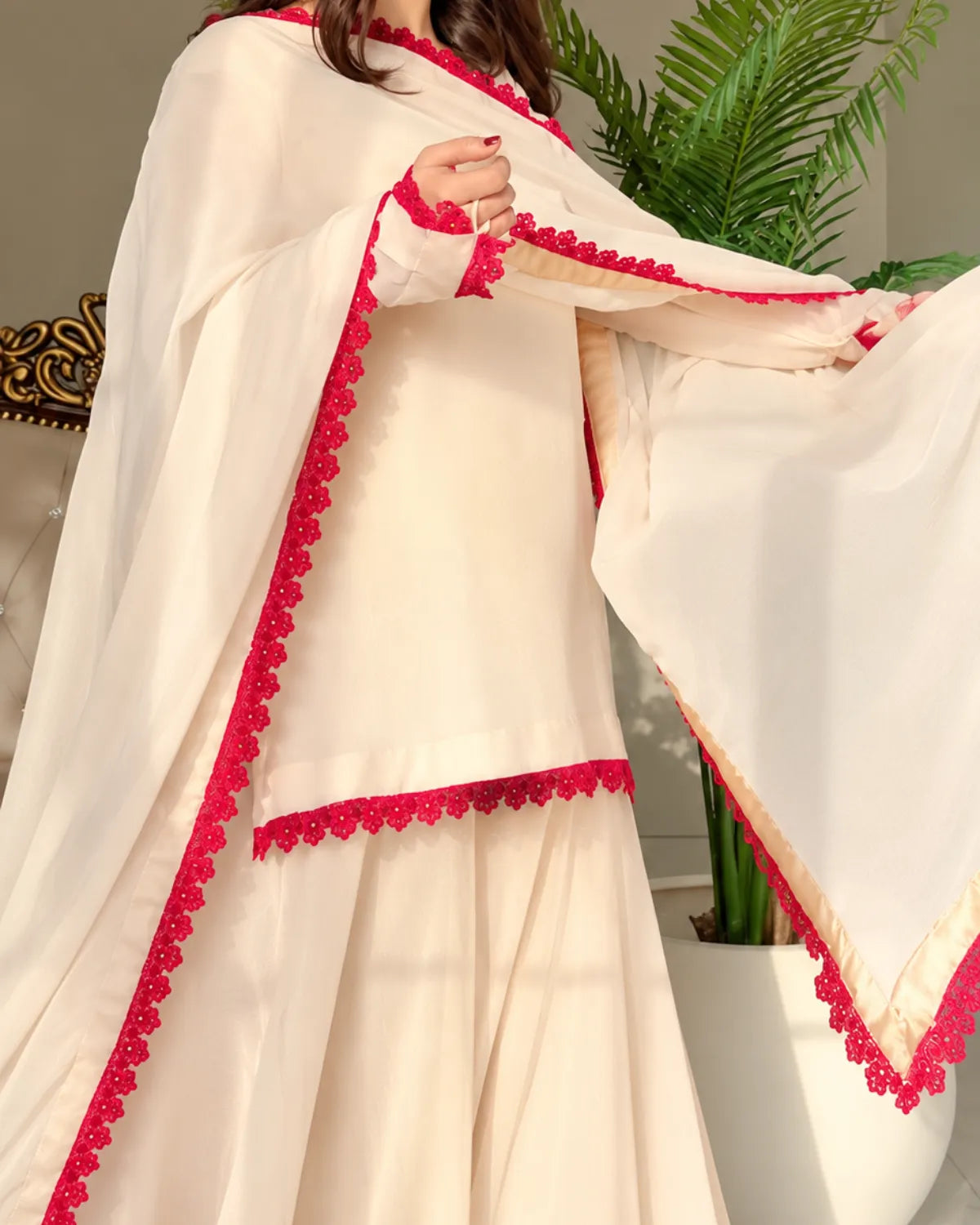A cream-colored traditional outfit with bright red scalloped lace trim, modeled in an indoor setting with a potted plant in the background.