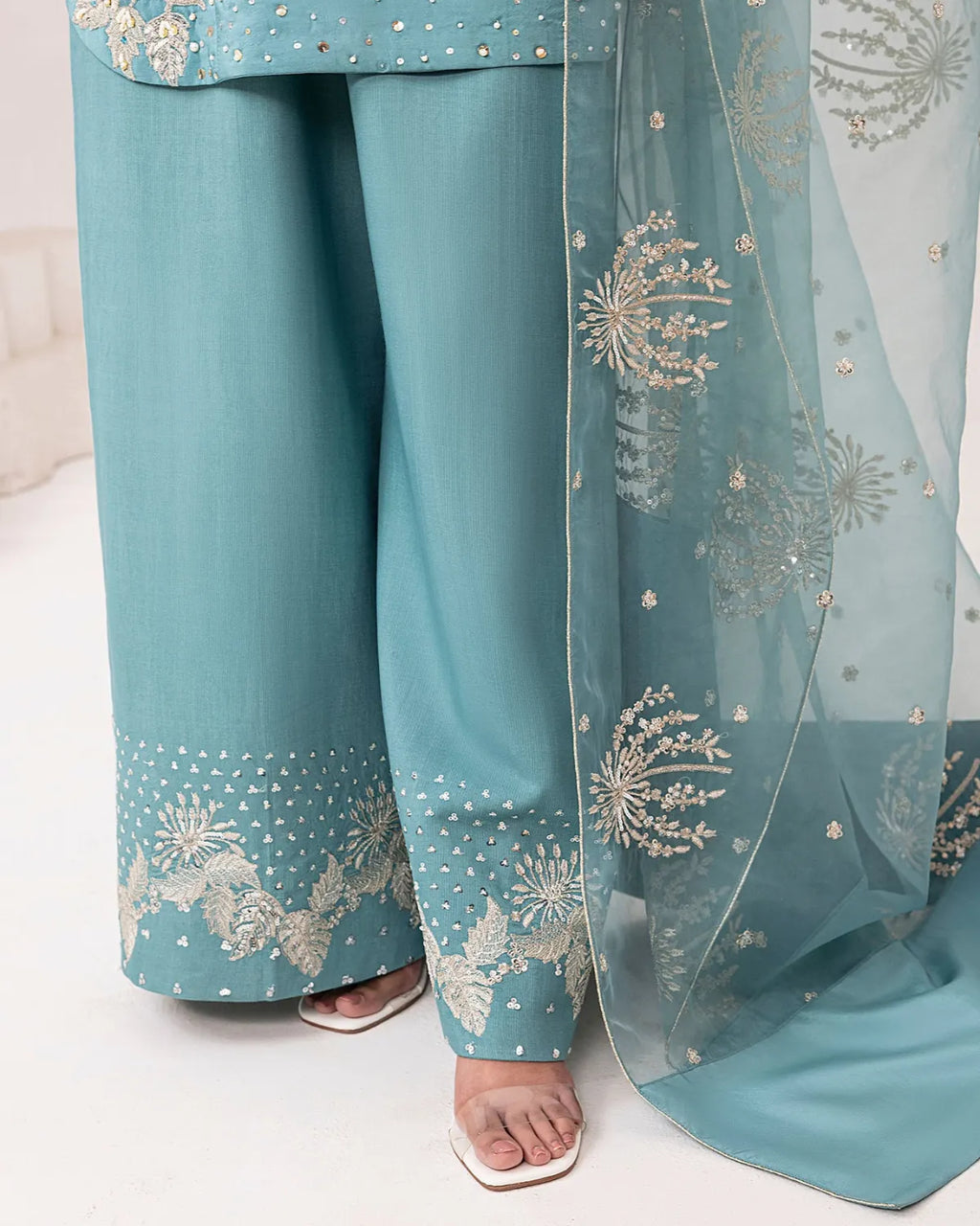 A close-up of a model wearing a teal blue traditional outfit with wide-leg pants, decorated with silver floral embroidery and dotted patterns, paired with a matching sheer dupatta.