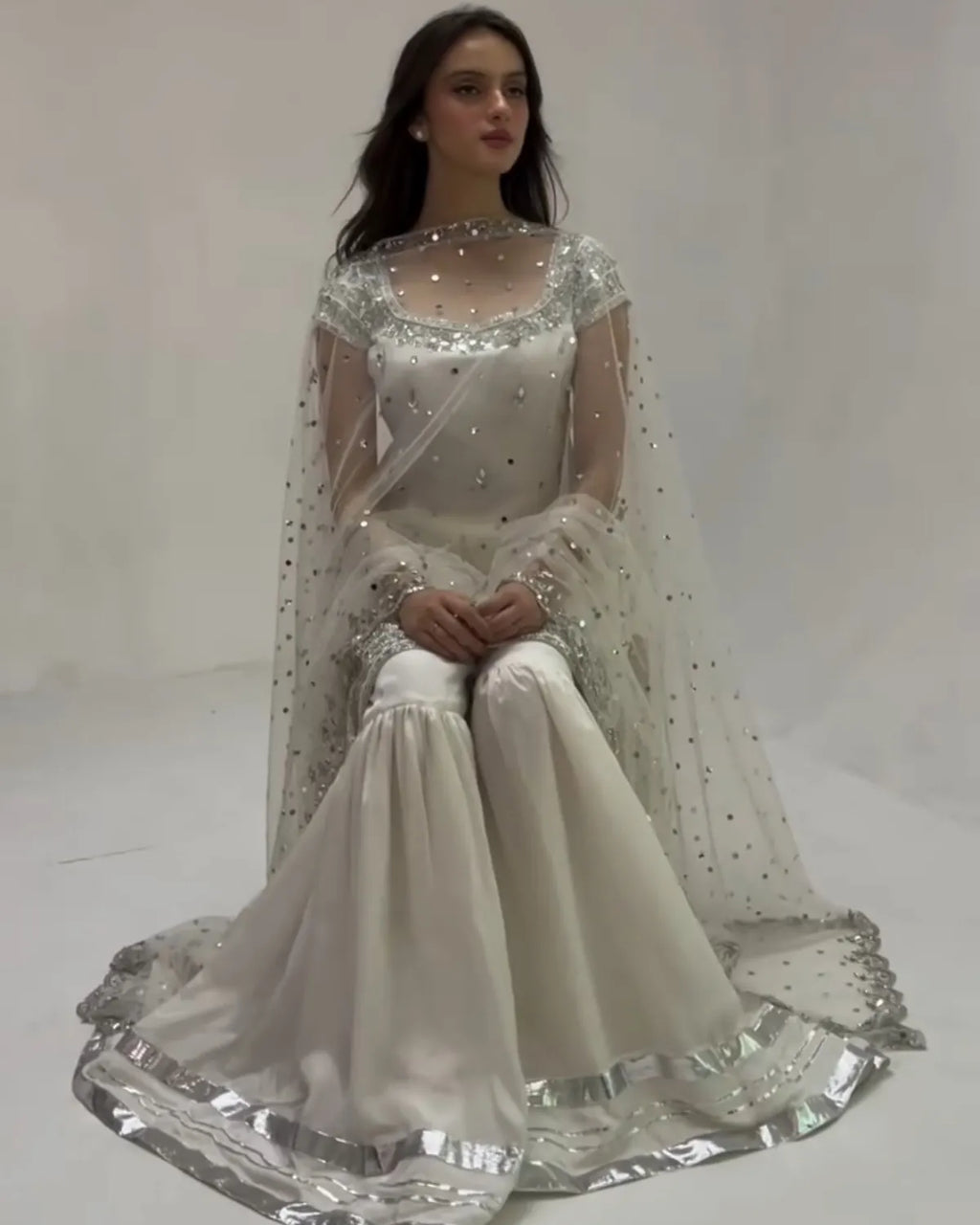 A woman seated in a studio wearing a formal white and silver embellished outfit with a sheer embroidered cape and wide flared pants.