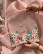 A close-up of a light pink embroidered fabric, likely a dupatta or scarf, held by hands to show its texture and detailed floral embroidery. The fabric features intricate embroidery with pink, green, and gold threads along a scalloped edge.