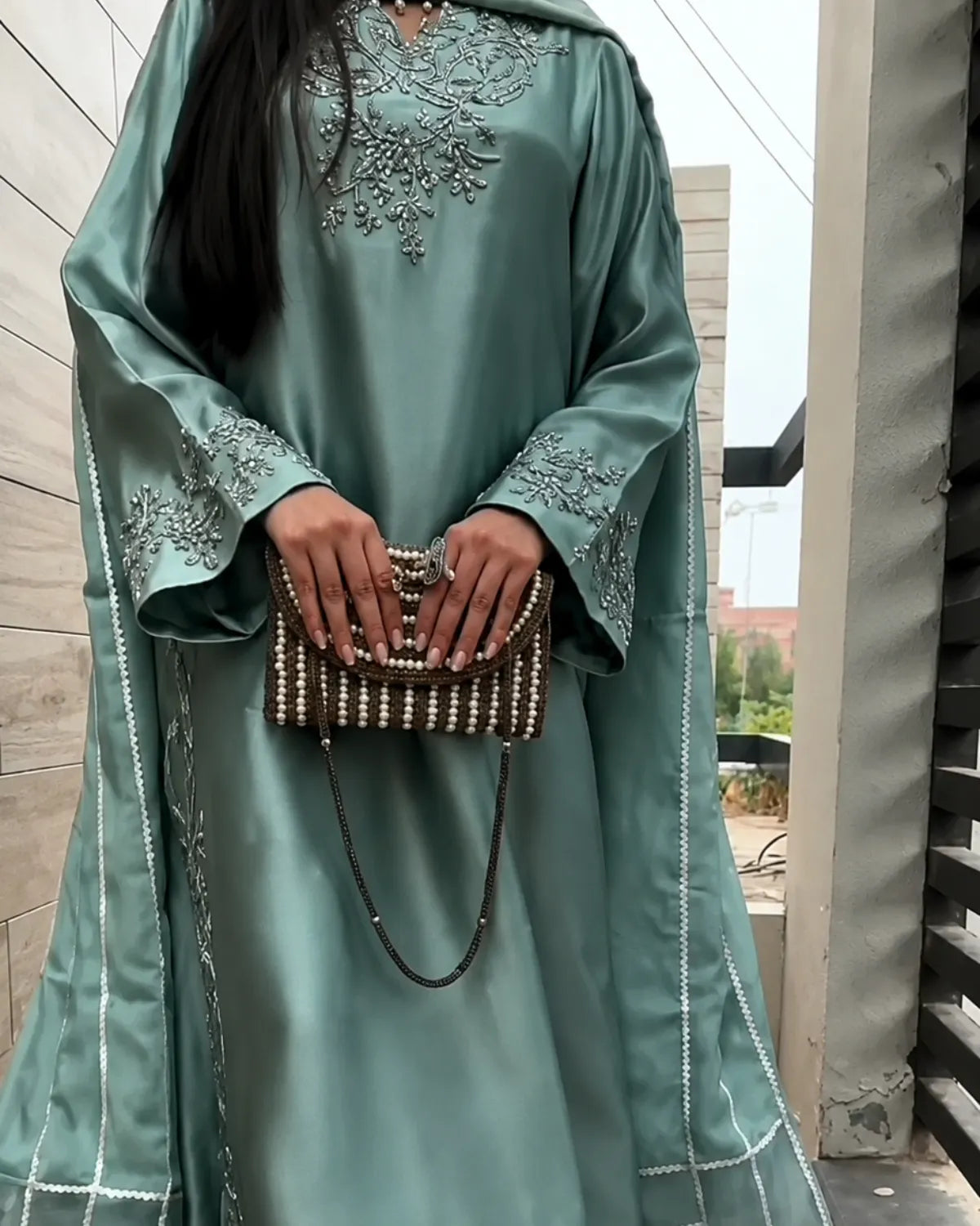 A woman wearing a flowing sage green abaya with silver embroidery on the chest and sleeves, holding a beaded brown and white clutch bag.